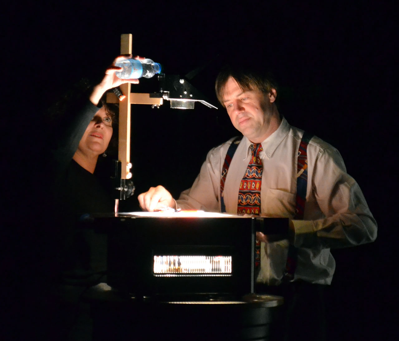 Extrait du spectacle : l'homme manipule des objets sur l'écran d'un rétroprojecteur, tandis que la femme verse de l'eau sur le miroir de l'appareil.