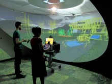 Un homme et une femme debout face à l'image projetée par le logiciel Hyve-3D. Ils agissent dans l'environnement virtuel au moyen de tablettes tactiles.